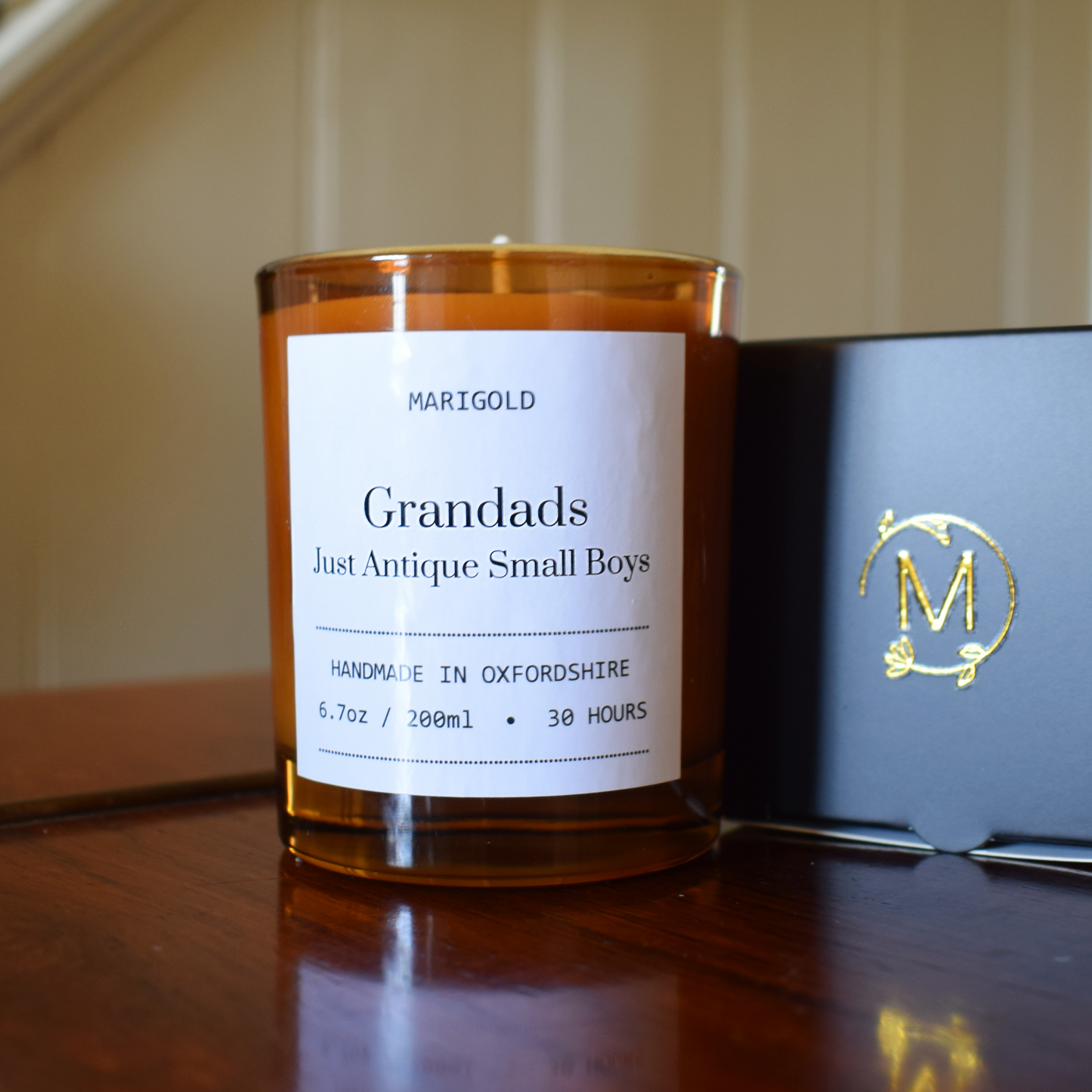 Candle with a label on a wooden surface next to a black box with a gold logo.