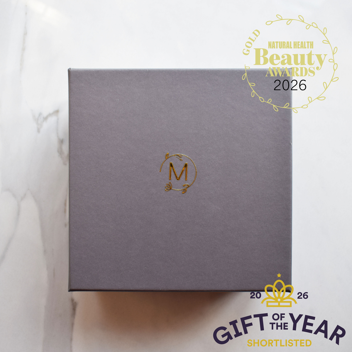A square dark gray gift box containing three Marigold skincare products with a gold monogram logo sits on a light marble surface. Award badges for Beauty Awards 2026 and Gift of the Year Shortlisted are overlaid in the corners.