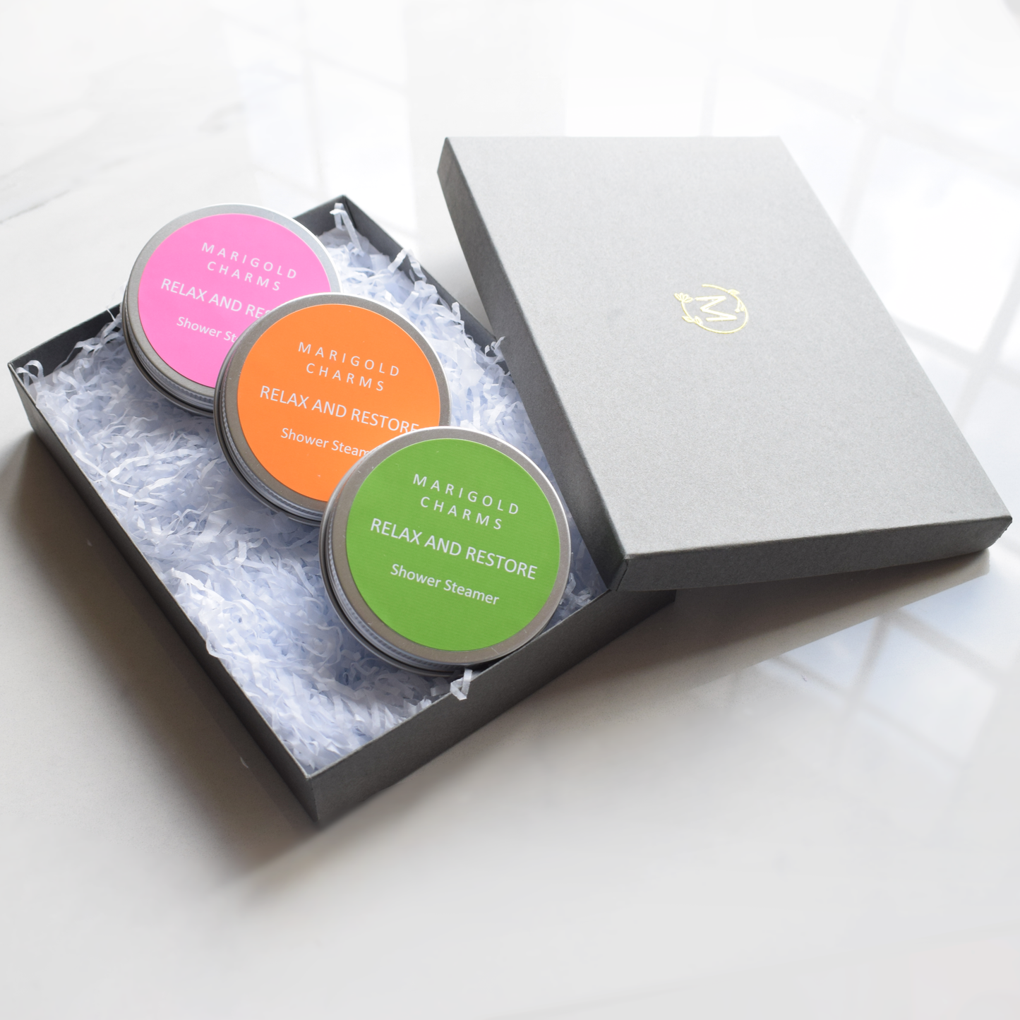 Three shower steamer containers in a box with 'Marigold Charms' branding on a white background.
