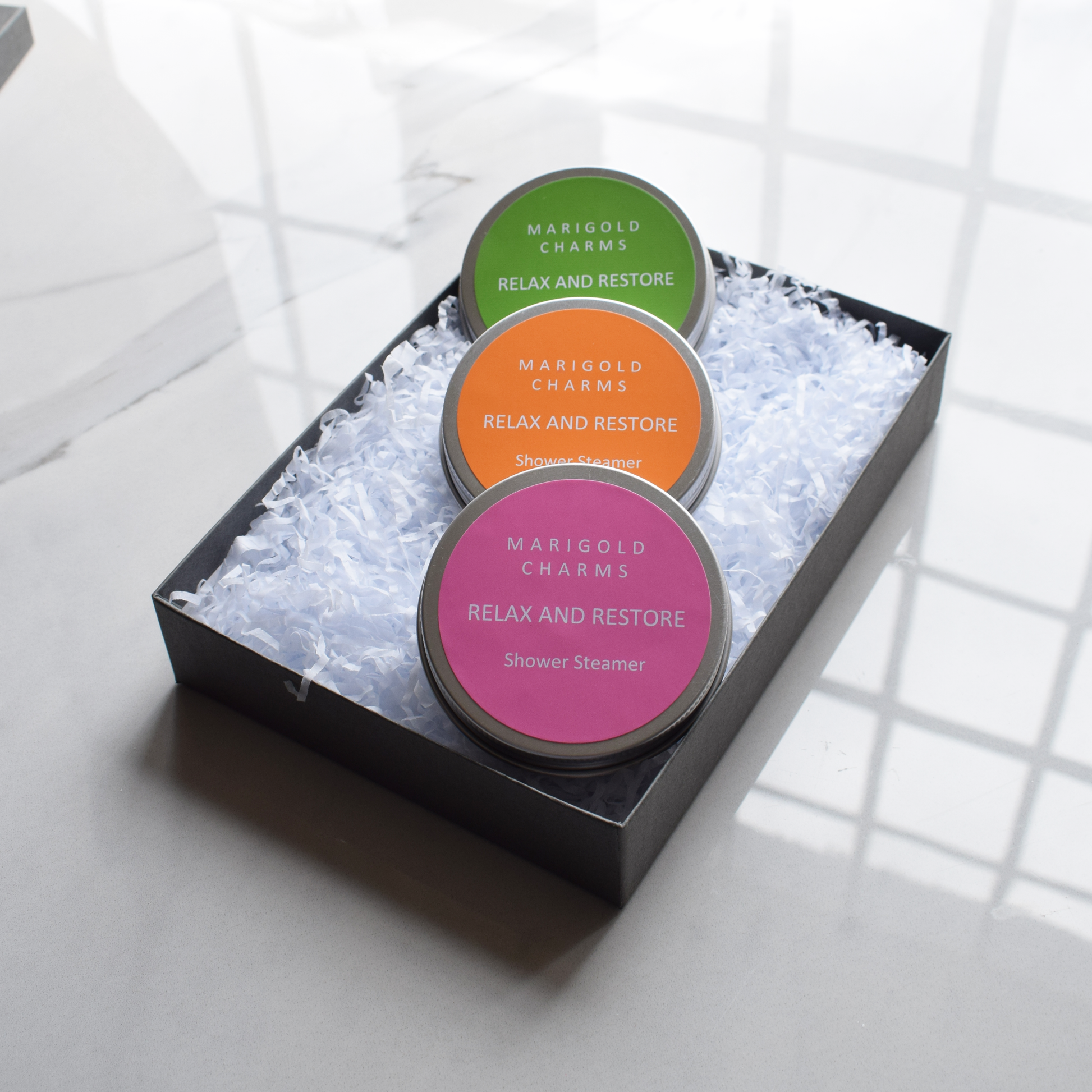 Three 'Marigold Charms' shower steamer containers in a black box on a light background