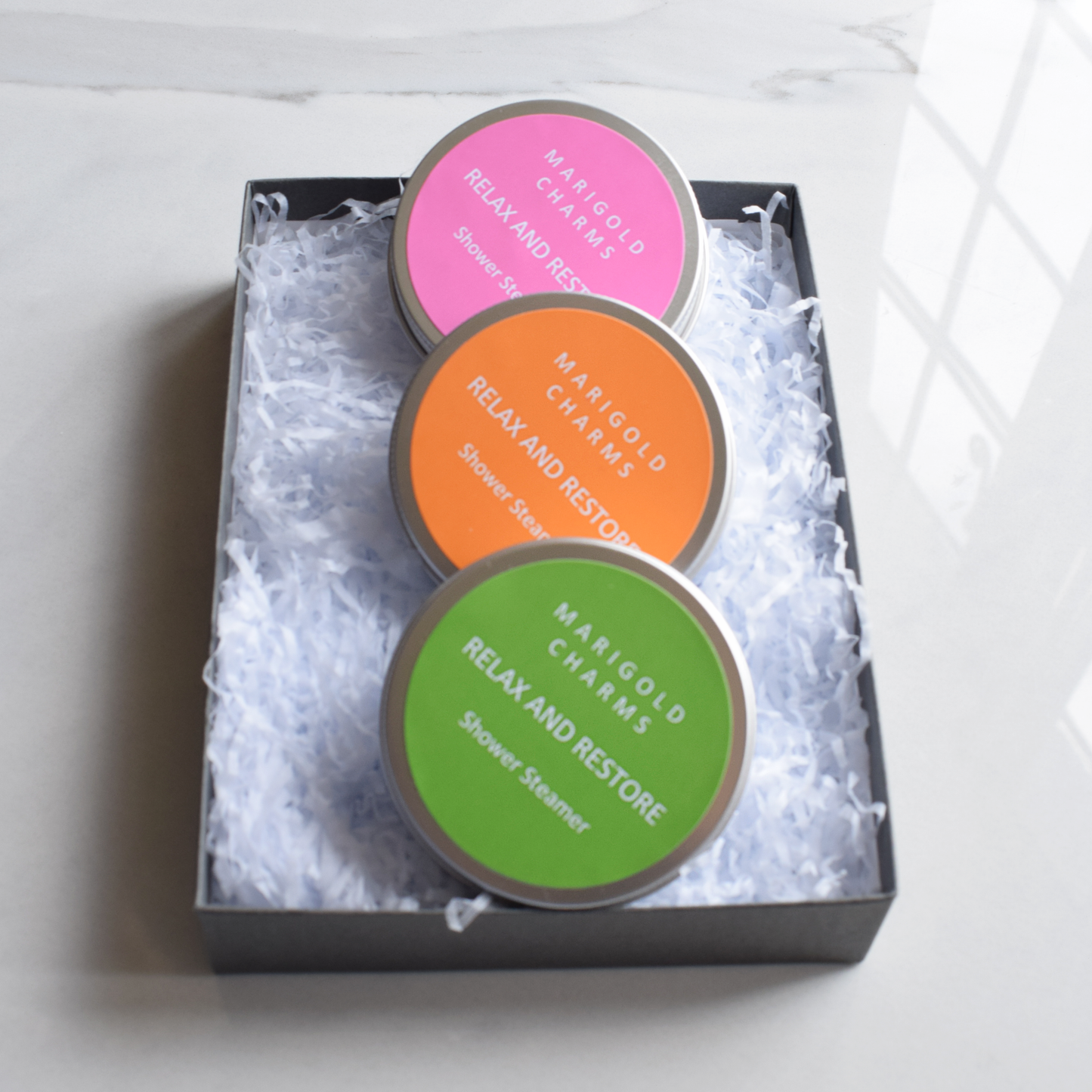 Three circular shower steamers in a box with colorful labels on a white background