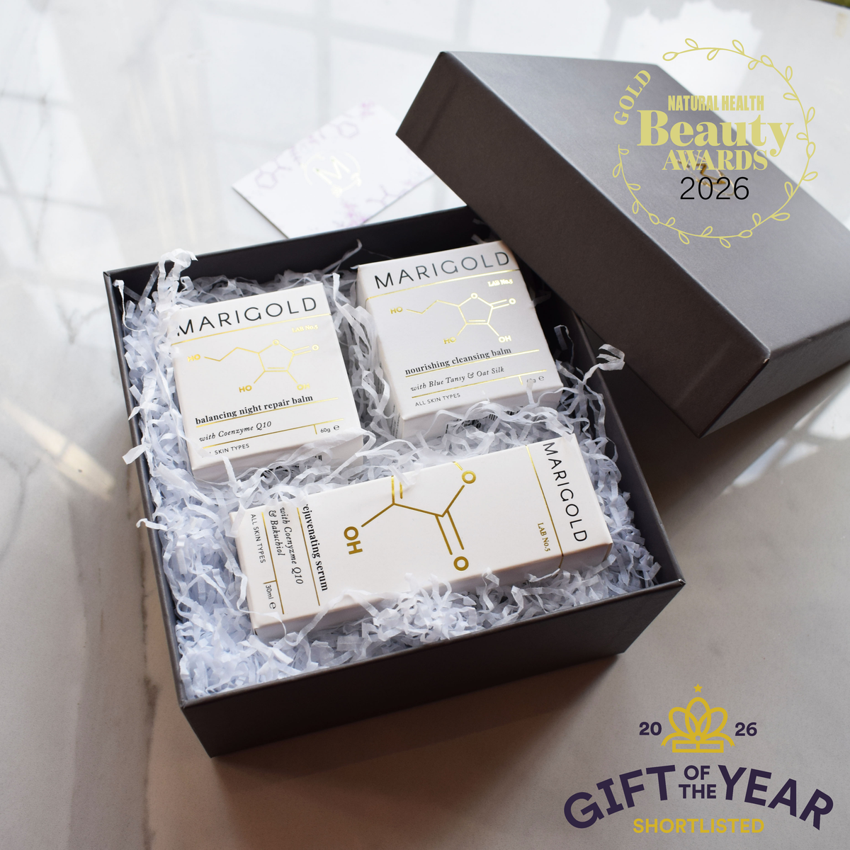 A luxury gift set of three MARIGOLD skincare products arranged in white crinkle paper inside a black box, with the black lid leaning against the background. A gold National Health Beauty Awards 2026 badge is on the lid, and a 'Gift of the Year 2026' badge is in the bottom right corner.