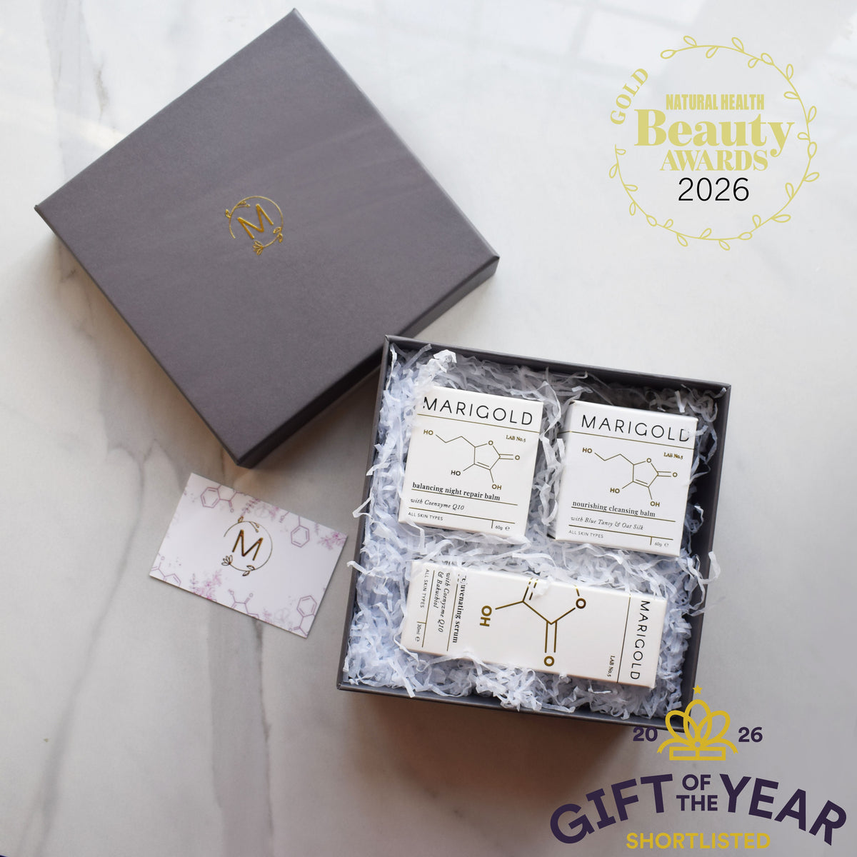 A luxury gift set from Marigold & Co. featuring a dark gray presentation box with a gold monogram logo, an open box filled with white shredded paper and three small branded soap bars, a small branded card, and a gold award badge overlay reading 'Gold Natural Health Beauty Awards 2026'.